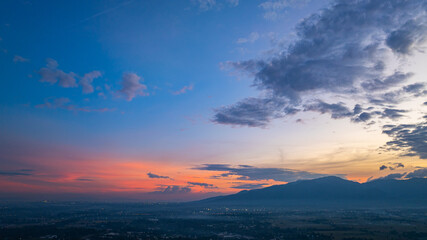 A breathtaking aerial panoramic view of a vibrant sunset illuminating the sky above a lush green hilltop with a small structure, surrounded by scattered clouds and distant landscapes.