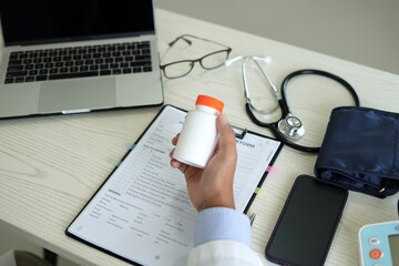 Doctor with stethoscope who is sitting in hospital clinic is holding an medicine bottle to assess...