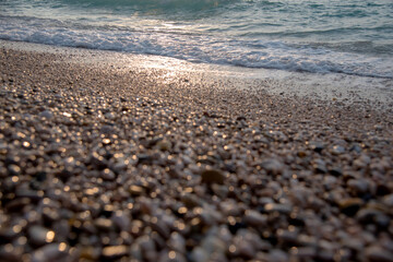 Close-up view of a beach with small, light brown and beige pebbles.  The focus is on the stones at the shoreline, with the water and waves slightly out of focus in the background. 