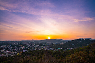 A scenic view of a town nestled among lush green mountains at sunset, with a temple structure perched on the hillside and a vibrant orange sky creating a warm, tranquil atmosphere.