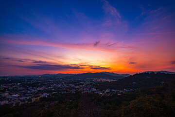 A scenic view of a town nestled among lush green mountains at sunset, with a temple structure perched on the hillside and a vibrant orange sky creating a warm, tranquil atmosphere.
