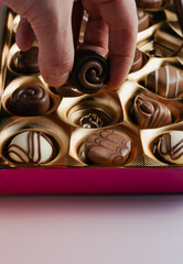 A human hand reaches into a gold box of assorted chocolates, picking up one of the delectable...