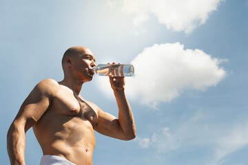 Athlete caucasian sun-tanned shirtless man drinking water against blue sky on outdoor sports ground. Concept water balance for hot season for active workout. Heat weather. Summer season.