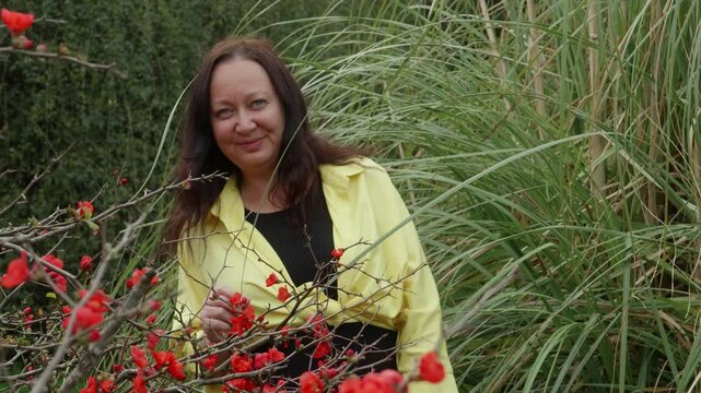Brunette woman wearing yellow overshirt and black undershirt gently caressing vibrant red flowers, experiencing peaceful moment in sunlit park during springtime leisure
