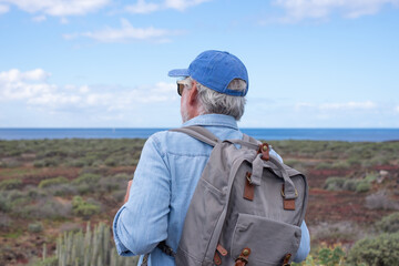 Back view of senior man with backpack in a trekking day in country footpath along the sea, enjoying healthy lifestyle and freedom, vacation or retirement concept
