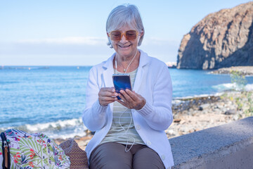 Smiling gray haired senior woman sitting relaxed near the beach using smartphone enjoying tech and social - retired lifestyle, horizon over the sea