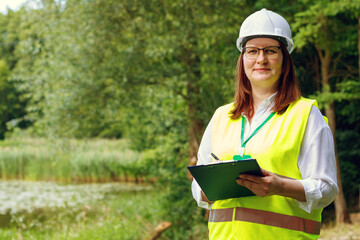Portrait of a European woman forestry inspector studying the condition of trees near water in the...