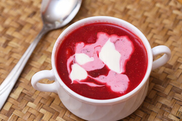 Traditional beetroot soup in white cup. Hot red borscht with sour cream swirl. Eastern European cuisine served with spoon. Rustic woven background and creamy topping.