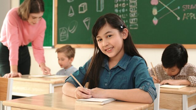 Asian girl writing test or classwork while looking at camera at classroom. Diverse happy student taking notes and teacher help caucasian boy doing test while sitting in front of blackboard. Pedagogy. - Powered by Adobe