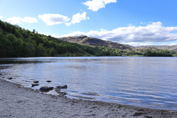 Shoreline of Grasmere Lake with calm water and surrounding hills under a partly cloudy blue sky. Lake District, England, UK.