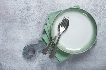 an empty plate and cutlery laid out, on a cutting board, with a green napkin, a gray table, top...