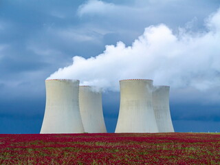Nuclear power plant cooling towers emitting steam over a red-flowered field under a dramatic cloudy...