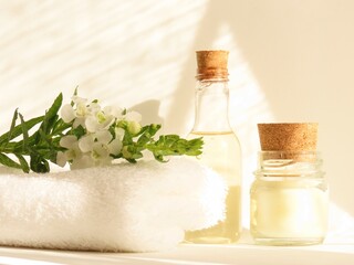 A small bottle of shower gel, a candle and a branch of white flowers and green leaves over a folded white towel with a sunlit white background. Spa still life.