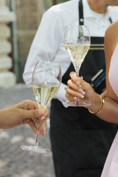 Close-up of two women clinking champagne glasses at an elegant outdoor event with a waiter in the background.