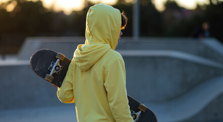 Person in yellow hoodie holding skateboard at skate park during golden hour sunset. Youth skateboarding culture and extreme sports lifestyle