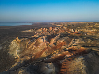 Aerial view follows the curving cliffs of Aktumsuk, revealing vibrant ochre, red, and beige bands. Former Aral Sea coastline stretches endlessly beyond, blending earth and sky at the horizon