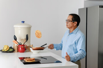 Confident elderly man flipping a pancake in the air while cooking breakfast in a modern kitchen. Concept of home cooking, active lifestyle, and joyful aging.