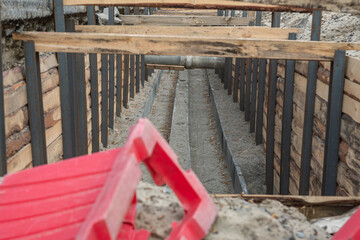 Long trench reinforced with wooden planks and metal bars, with part of a red plastic fence visible in the foreground.