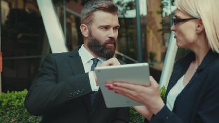 Serious business man and woman talking standing and use tablet computer together at outdoor handsome internet technology team corporate executive city outside urban young teamwork worker slow motion - Powered by Adobe