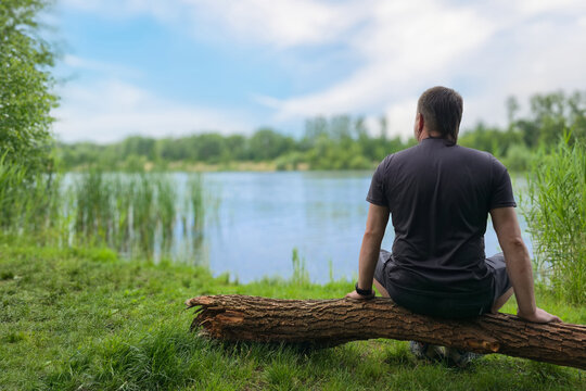Young man sitting by lake in nature, seen from behind, relaxing in solitude. Back view of alone young male in casual hiking clothes sitting on log near calm forest lake or river. mental balance