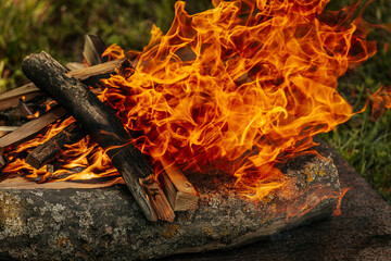 Bright orange flames rise from burning firewood arranged on a stone surface in an outdoor setting,...