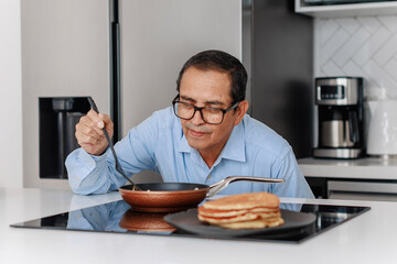 Elderly man in blue shirt checking pancake in frying pan with spatula in modern kitchen. Stack of...