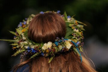 Naklejka premium Close-up of a flower crown made of colorful wildflowers and greenery on a woman's brown hair, viewed from behind during a traditional outdoor celebration.