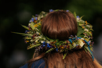 Naklejka premium Close-up of a flower crown made of colorful wildflowers and greenery on a woman's brown hair, viewed from behind during a traditional outdoor celebration.