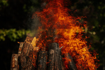 Tall bonfire burning intensely with orange flames reaching high, surrounded by rocks and trees...