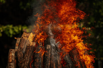 Tall bonfire burning intensely with orange flames reaching high, surrounded by rocks and trees...
