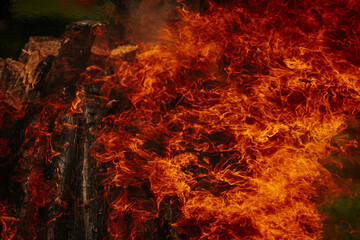 Tall bonfire burning intensely with orange flames reaching high, surrounded by rocks and trees...