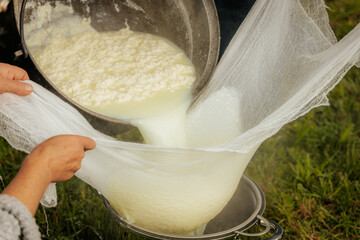 Curds and whey are being poured from a large pot into cheesecloth to drain over a colander during...