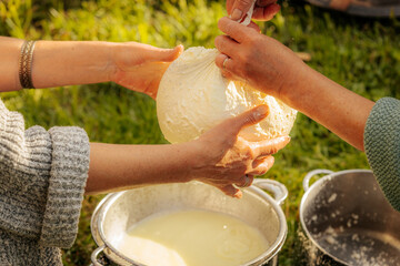 Close-up of hands holding fresh cheese wrapped in cloth, draining whey into a strainer, part of...