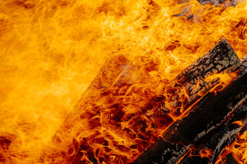 Intense orange flames engulf burning firewood in a large bonfire, with vivid textures of charred...