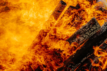 Intense orange flames engulf burning firewood in a large bonfire, with vivid textures of charred...