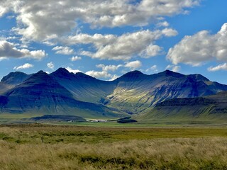 Layered mountains in East Iceland with light and shadow play over grassy plains and a distant farm...
