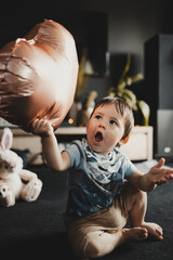 Surprised child in a gray T-shirt and beige shorts, holding a heart-shaped copper balloon, sitting on a dark floor in the comfort of home, with a blurred background
