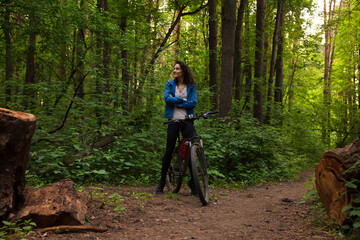 Obraz premium Beauty smiling sport free woman biking on bicycle on path with logs in green woods in a summer forest. Strong body, legs girl outdoors. Female walking in park in sporty mountain bike.