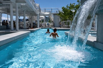 Two boys are seen enjoying themselves in a vibrant swimming pool, with a beautiful waterfall feature, capturing the joy of leisure activities on a sunny summer day.