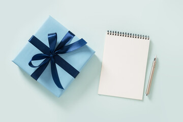 Blue gift box, white blank, spiral notebook and pen on pastel blue background. Invitation. Flat lay. View from above. Copy space.