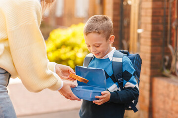 Mother gives happy boy with backpack a healthy lunchbox before school, back to school morning, outdoors
