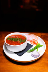 Traditional red borscht soup with fresh herbs, served in a white bowl with sliced ham, tomatoes, green onion, garlic, and red onion. Classic Eastern European cuisine