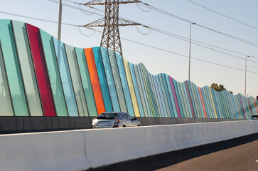 A colorful sound barrier panel installation along a highway, featuring a wave-like design composed of multicolored vertical glass and acrylic panels. A noise-reduction wall of urban infrastructure.