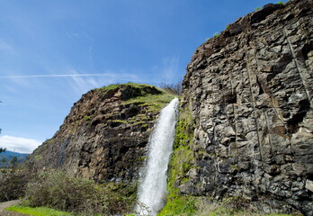 Rowland Lake Falls with wide swath of water crashing down between rough rocky walls such that the spray increases the local humidity enough to allow moss and other green foliage to grow on the cliff.