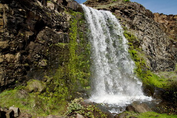 Rowland Lake Falls with wide swath of water crashing down between rough rocky walls such that the spray increases the local humidity enough to allow moss and other green foliage to grow on the cliff.