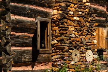 A side of a log cabin with a stone chimney and open window with a shutter