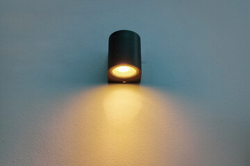 A modern, cylindrical dark grey or black wall lamp illuminates a textured white or light-colored wall with a warm, downward-facing glow.