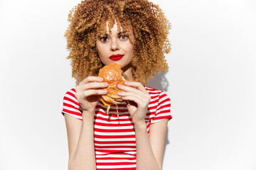 Beautiful young woman with curly hair enjoying a delicious burger, wearing a red and white striped shirt, vibrant colors and playful expression ideal for food marketing