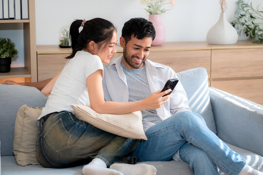 Young asian couple relax sitting on sofa looking smartphone together in living room at home, cozy lifestyle and bonding, relationship and leisure, sharing and enjoy digital content with happy.