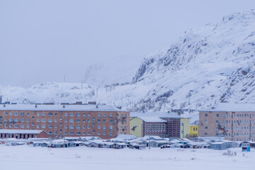 Snow-covered residential buildings in a remote area, surrounded by majestic mountains and a winter...